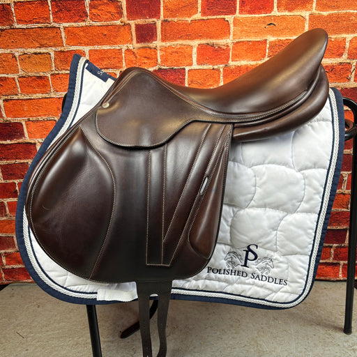A Butet 'C' Monoflap Jumping Saddle made of medium brown leather, displayed on a white saddle pad with black and blue trim, against a brick wall background.